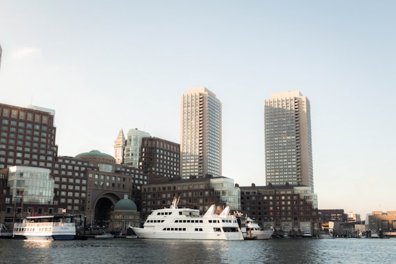 A waterfront cityscape featuring two prominent high-rise buildings surrounded by other modern architecture. Several boats and yachts are docked along the waterside, with clear skies in the background.