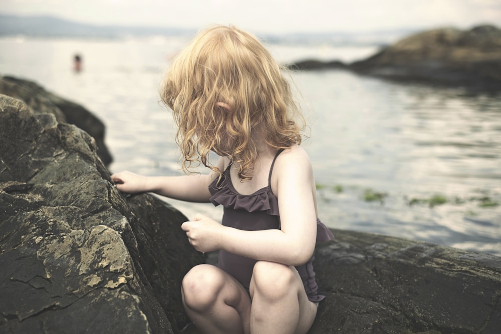 girl in black tank top sitting on rock near body of water during daytime