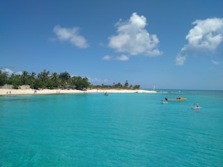 A tranquil beach scene with clear turquoise waters and a sandy shore lined with lush green palm trees. Several people are enjoying leisure activities, including kayaking and sunbathing. Fluffy white clouds dot the bright blue sky, enhancing the serene atmosphere.
