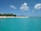 A tranquil beach scene with clear turquoise waters and a sandy shore lined with lush green palm trees. Several people are enjoying leisure activities, including kayaking and sunbathing. Fluffy white clouds dot the bright blue sky, enhancing the serene atmosphere.
