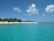 A tranquil beach scene with clear turquoise waters and a sandy shore lined with lush green palm trees. Several people are enjoying leisure activities, including kayaking and sunbathing. Fluffy white clouds dot the bright blue sky, enhancing the serene atmosphere.
