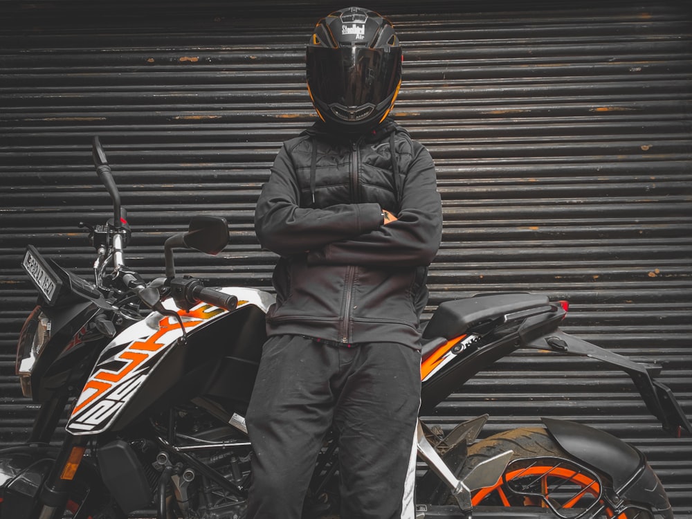 검은 색과 주황색 오토바이 헬멧과 회색 재킷을 입은 남자가 검은 색과 주황색 스포츠 옆에 서 있다