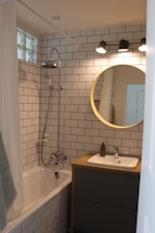A modern bathroom with white subway tile walls, a bathtub with a shower, a circular mirror above a wooden vanity, and three overhead lights. The sink has a soap dispenser, and a towel hangs on the side of the bathtub.