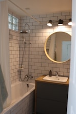 A modern bathroom with white subway tile walls, a bathtub with a shower, a circular mirror above a wooden vanity, and three overhead lights. The sink has a soap dispenser, and a towel hangs on the side of the bathtub.