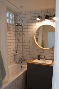 A modern bathroom with white subway tile walls, a bathtub with a shower, a circular mirror above a wooden vanity, and three overhead lights. The sink has a soap dispenser, and a towel hangs on the side of the bathtub.