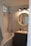 A modern bathroom with white subway tile walls, a bathtub with a shower, a circular mirror above a wooden vanity, and three overhead lights. The sink has a soap dispenser, and a towel hangs on the side of the bathtub.