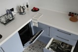 A modern kitchen counter with a white surface, featuring a small kettle, a dish rack with cups and a plate, two red apples on a black tray, and spice jars. A tea towel is draped over the edge. Below the counter are open dishwashers and drawers containing metallic pots and pans.