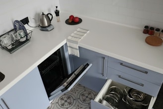 A modern kitchen counter with a white surface, featuring a small kettle, a dish rack with cups and a plate, two red apples on a black tray, and spice jars. A tea towel is draped over the edge. Below the counter are open dishwashers and drawers containing metallic pots and pans.