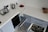 A modern kitchen counter with a white surface, featuring a small kettle, a dish rack with cups and a plate, two red apples on a black tray, and spice jars. A tea towel is draped over the edge. Below the counter are open dishwashers and drawers containing metallic pots and pans.