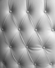 A detailed image of tufted upholstery with a diamond pattern and buttons securing the fabric at regular intervals. The texture of the material is clearly visible, with a soft sheen indicating high-quality leather or a similar material.