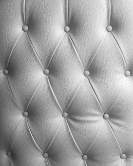 A detailed image of tufted upholstery with a diamond pattern and buttons securing the fabric at regular intervals. The texture of the material is clearly visible, with a soft sheen indicating high-quality leather or a similar material.
