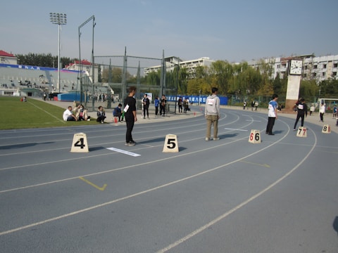 A running track with several lanes marked by numbers. Some individuals are standing on the track while others are seated near the sides. The scene includes a clock tower on the right side and a stadium area with seats on the left. There are other people scattered around and buildings in the background under a clear sky.