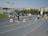 A running track with several lanes marked by numbers. Some individuals are standing on the track while others are seated near the sides. The scene includes a clock tower on the right side and a stadium area with seats on the left. There are other people scattered around and buildings in the background under a clear sky.