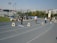 A running track with several lanes marked by numbers. Some individuals are standing on the track while others are seated near the sides. The scene includes a clock tower on the right side and a stadium area with seats on the left. There are other people scattered around and buildings in the background under a clear sky.