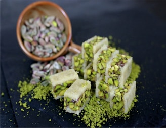 A collection of white nougat pieces densely packed with vibrant green pistachio nuts is arranged on a dark surface. Nearby, an overturned wooden bowl spills out more pistachios, adding to the scattered green pieces around the sweets.