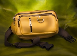 A yellow leather waist bag with multiple zippered compartments and a metallic ring, placed on a green background. It has a black adjustable strap with a plastic buckle.