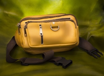 A yellow leather waist bag with multiple zippered compartments and a metallic ring, placed on a green background. It has a black adjustable strap with a plastic buckle.
