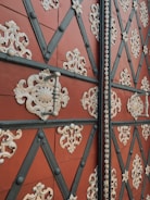 An ornate door with a geometric pattern of black diagonal lines forming diamond shapes. These diamonds are adorned with intricate cream-colored floral and scrollwork metal embellishments. The base color of the door is a rich, earthy red, with metallic handles and studs adding texture.