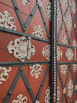 An ornate door with a geometric pattern of black diagonal lines forming diamond shapes. These diamonds are adorned with intricate cream-colored floral and scrollwork metal embellishments. The base color of the door is a rich, earthy red, with metallic handles and studs adding texture.