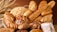 A basket filled with a variety of freshly baked bread, including baguettes, round loaves, and rolls. The bread features different textures and crust patterns, with some topped with seeds. To the left, wheat stalks add an organic touch to the arrangement.