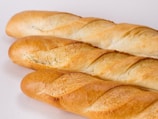 Three fresh baguettes with a golden crust are arranged on a plain white background. The bread loaves have a crisp, slightly uneven texture and a lightly browned exterior with scoring marks.