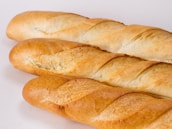 Three fresh baguettes with a golden crust are arranged on a plain white background. The bread loaves have a crisp, slightly uneven texture and a lightly browned exterior with scoring marks.