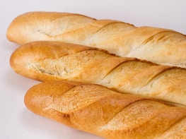 Three fresh baguettes with a golden crust are arranged on a plain white background. The bread loaves have a crisp, slightly uneven texture and a lightly browned exterior with scoring marks.