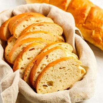 A basket lined with a beige cloth holds several slices of fresh, crusty bread. The slices are arranged neatly in an overlapping pattern, showcasing their golden-brown crust and soft, airy interior. In the background, part of an unsliced loaf is visible.