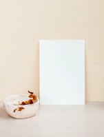 A minimalist composition featuring a blank white sheet of paper propped upright against a light beige background. In the foreground, there is a small ceramic bowl with a rough texture, containing a few dried leaves or petals, adding a touch of organic contrast.