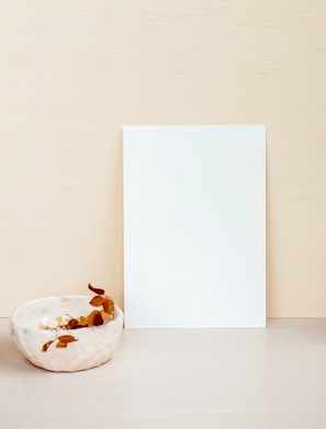 A minimalist composition featuring a blank white sheet of paper propped upright against a light beige background. In the foreground, there is a small ceramic bowl with a rough texture, containing a few dried leaves or petals, adding a touch of organic contrast.