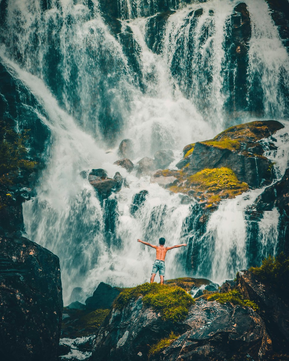 man in red shirt standing on rock near waterfalls during daytime