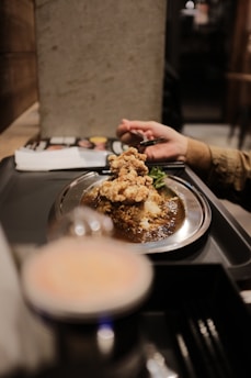 A person holding a spoon is about to eat a dish featuring crispy fried chicken atop a mound of rice covered in thick, dark curry sauce on a metal plate. The setting appears to be casual dining, with a magazine visible in the background.