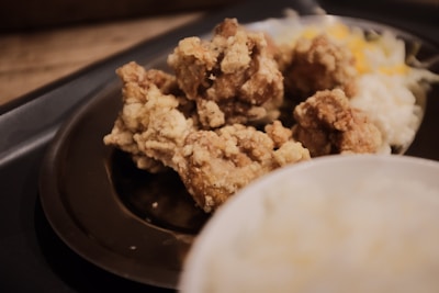 A close-up view of fried chicken pieces on a dark plate, accompanied by a serving of shredded cheese and a portion of mashed potatoes. The foreground features a blurred white object, possibly a bowl or plate.