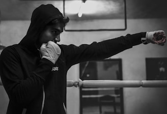 A person wearing a hooded sweater and boxing gloves is practicing a punch inside a boxing ring. The environment appears to be a gym or training area with minimal lighting.