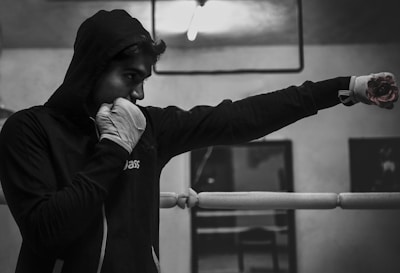 A person wearing a hooded sweater and boxing gloves is practicing a punch inside a boxing ring. The environment appears to be a gym or training area with minimal lighting.