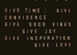 A series of motivational phrases are displayed in neon-style text against a dark background. The phrases include 'give time', 'give confidence', 'give good vibes', 'give joy', 'give inspiration', and 'give love'. The words are arranged in a structured, grid-like format, creating a visually impactful and positive message.
