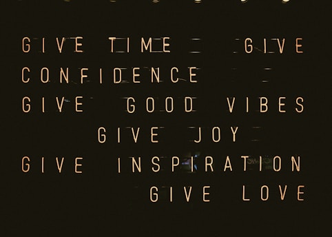 A series of motivational phrases are displayed in neon-style text against a dark background. The phrases include 'give time', 'give confidence', 'give good vibes', 'give joy', 'give inspiration', and 'give love'. The words are arranged in a structured, grid-like format, creating a visually impactful and positive message.
