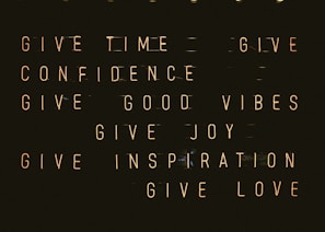 A series of motivational phrases are displayed in neon-style text against a dark background. The phrases include 'give time', 'give confidence', 'give good vibes', 'give joy', 'give inspiration', and 'give love'. The words are arranged in a structured, grid-like format, creating a visually impactful and positive message.