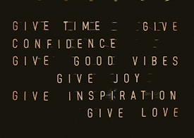 A series of motivational phrases are displayed in neon-style text against a dark background. The phrases include 'give time', 'give confidence', 'give good vibes', 'give joy', 'give inspiration', and 'give love'. The words are arranged in a structured, grid-like format, creating a visually impactful and positive message.