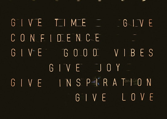A series of motivational phrases are displayed in neon-style text against a dark background. The phrases include 'give time', 'give confidence', 'give good vibes', 'give joy', 'give inspiration', and 'give love'. The words are arranged in a structured, grid-like format, creating a visually impactful and positive message.