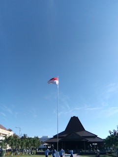A tall flagpole with a red and white flag is planted in front of a traditional, large building with a triangular roof. The sky above is clear and blue. Several people dressed in blue uniforms are standing at the base of the flagpole. There are well-maintained gardens and trees surrounding the area.