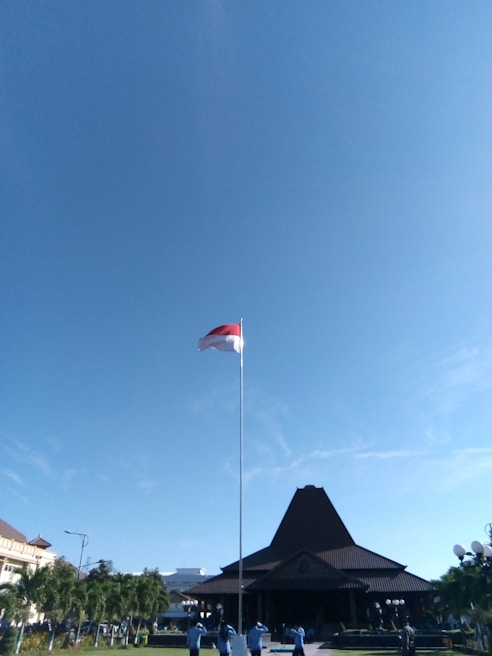 A tall flagpole with a red and white flag is planted in front of a traditional, large building with a triangular roof. The sky above is clear and blue. Several people dressed in blue uniforms are standing at the base of the flagpole. There are well-maintained gardens and trees surrounding the area.