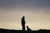 man in black jacket standing beside white dog on green grass field during daytime
