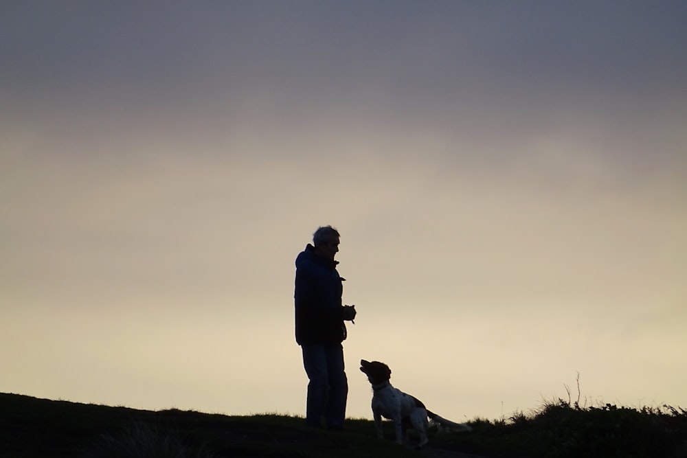 man in black jacket standing beside white dog on green grass field during daytime