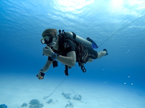 A scuba diver swims underwater, equipped with breathing apparatus and diving gear. The diver is surrounded by a vast expanse of clear blue water with the ocean floor visible below.