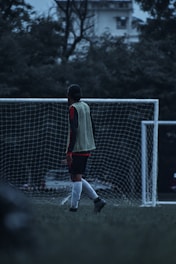 A soccer player stands on a field in front of a goal post, wearing a green training vest over a red and black long-sleeved shirt. The background is filled with trees, and the overall scene appears to be during twilight or early morning due to low light conditions.
