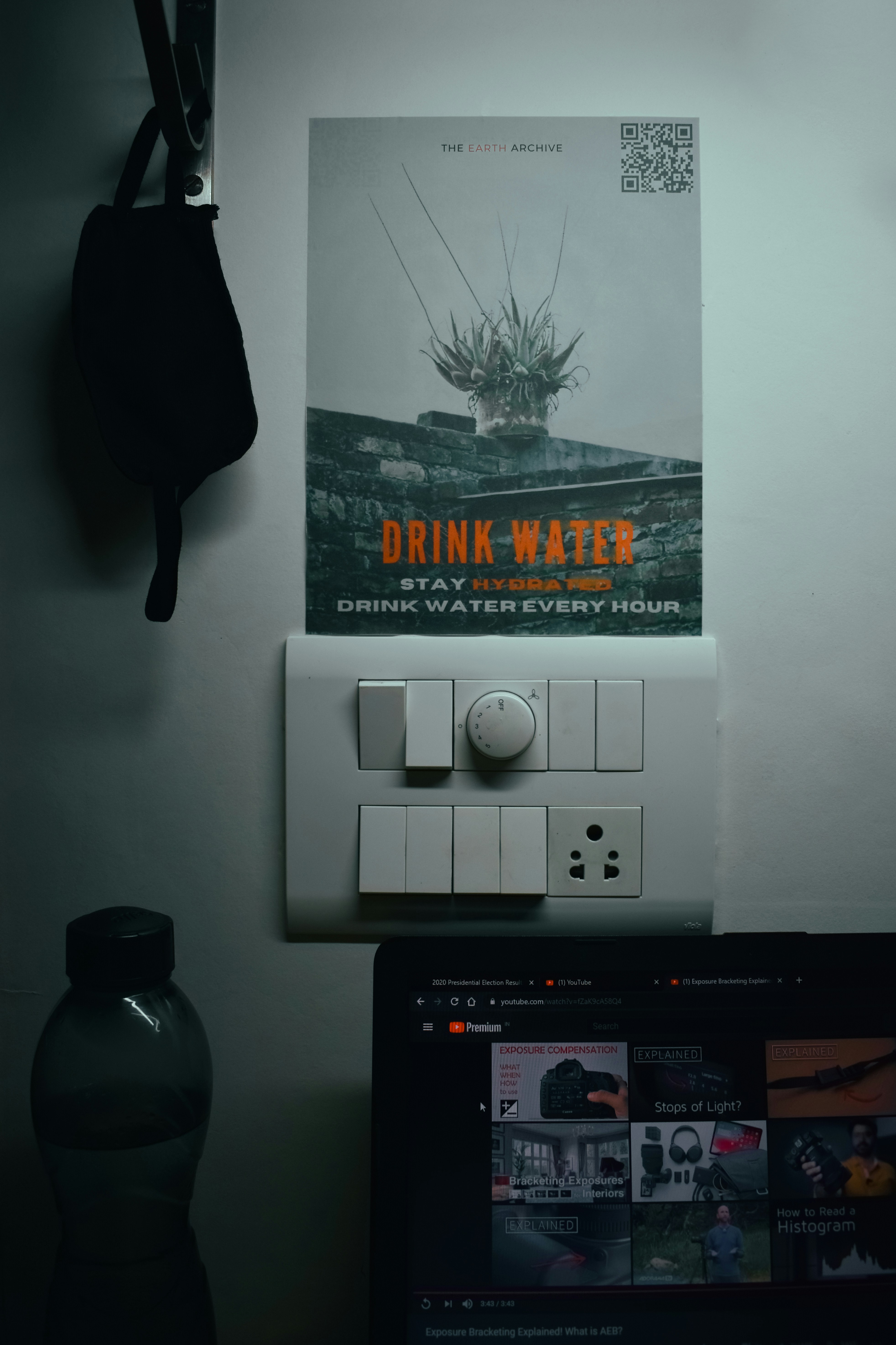 An open laptop with YouTube running, with a water bottle beside it and a mask hanging up. A poster advising to drink water is stuck on the wall.