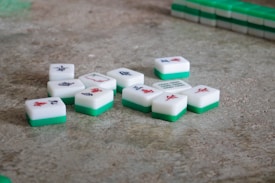 Several Mahjong tiles are scattered on a textured surface. The tiles have a white top with various Chinese characters and symbols in red and blue, and a green base. In the background, additional tiles are stacked neatly.