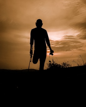 A silhouette of a person standing on one leg, using crutches, against a warm, dramatic sunset or sunrise sky. The surrounding landscape is slightly visible with outlines of plants and hills.