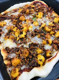 A close-up view of a homemade pizza topped with various ingredients such as fried cauliflower, spices, and bits of herbs on a thin crust. The edges appear slightly crisped, and the toppings are evenly spread across the surface, giving it a hearty and rustic appearance.